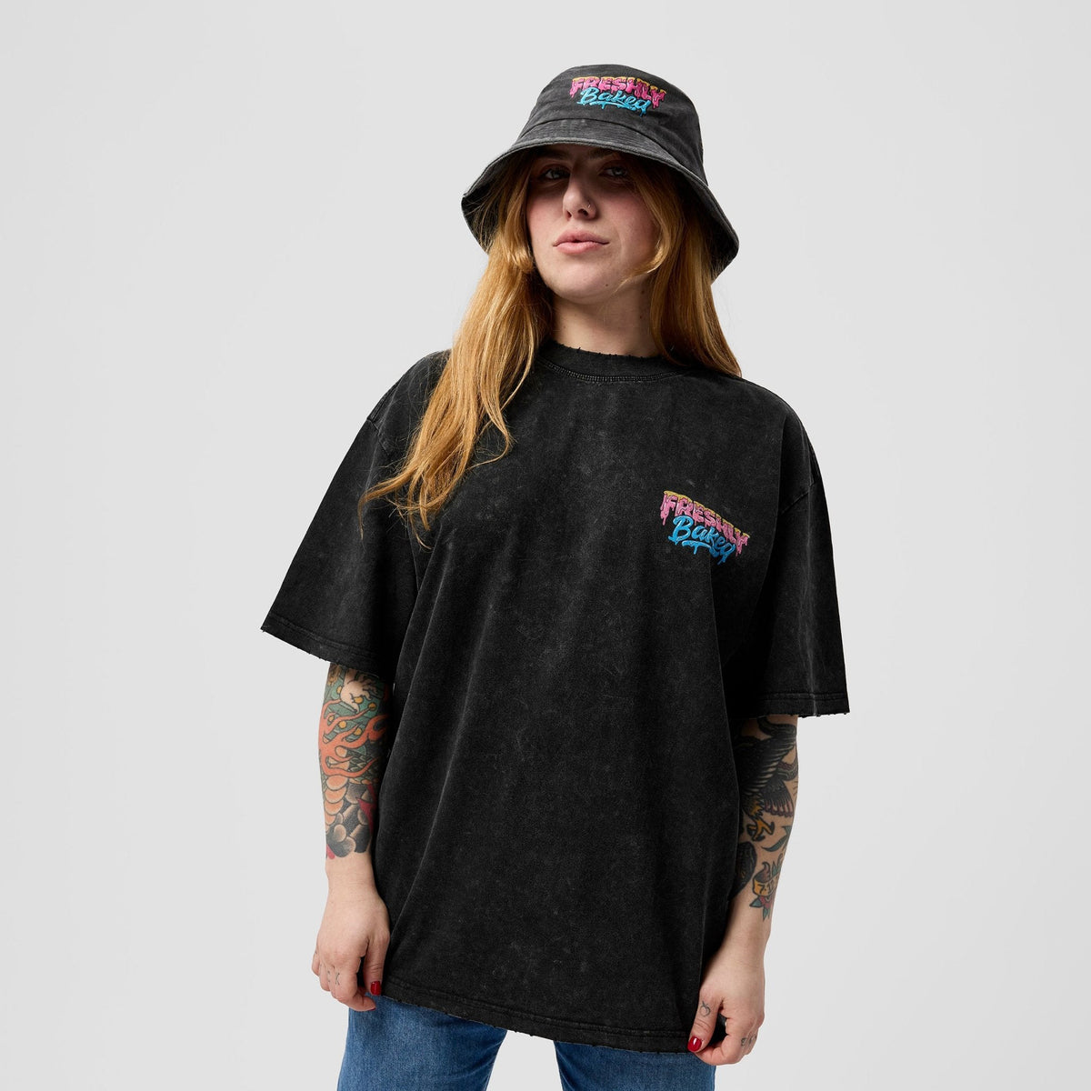A woman wearing a black acid-washed bucket hat and an oversized black acid-washed t-shirt with a colorful 'Freshly Picked' logo on the chest, paired with blue jeans, against a light gray background.