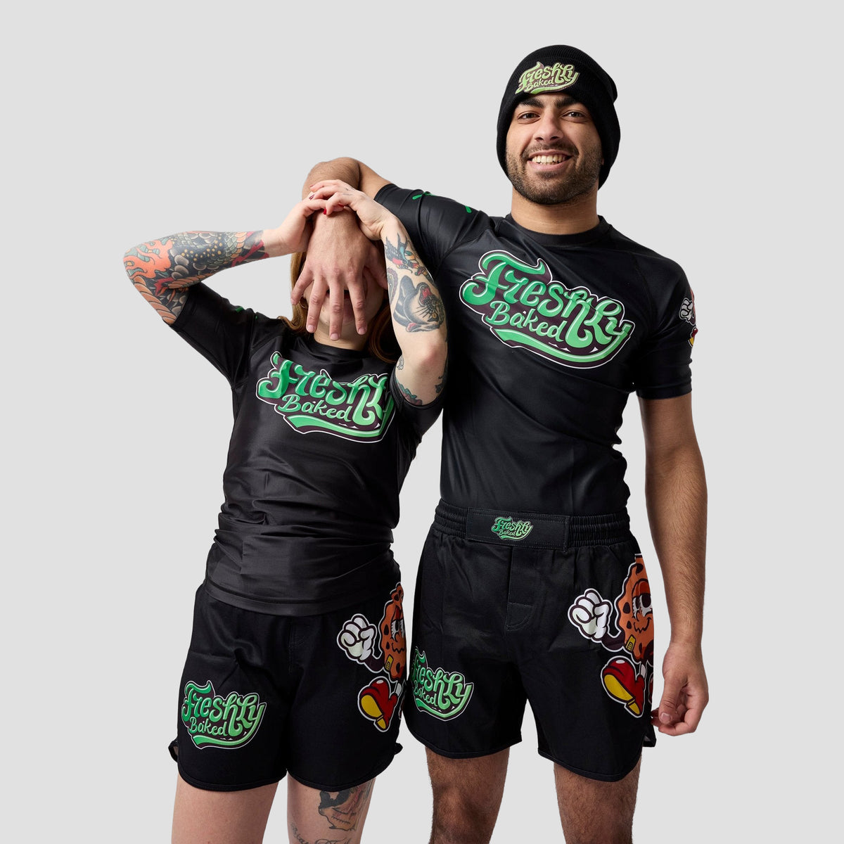 Two models wearing matching black Freshly Baked apparel, including a short-sleeve t-shirt, shorts, and a beanie, posed against a solid light gray background.