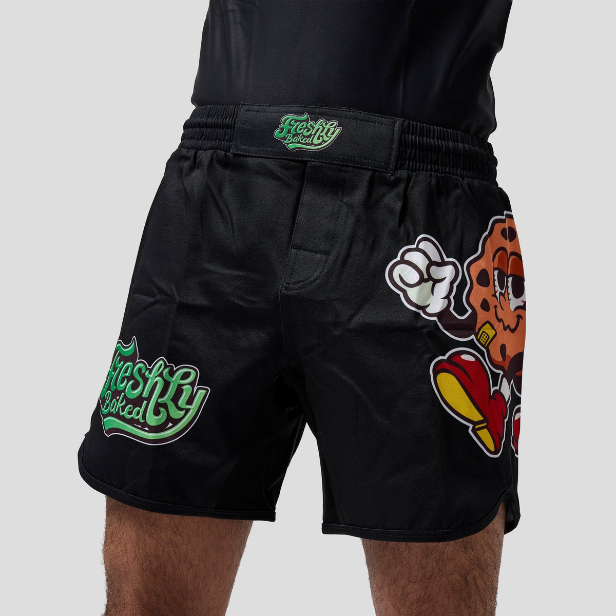 Black athletic shorts worn by a model, featuring green 'Freshly Baked' branding on the waistband and left leg, along with a large cartoon cookie graphic on the right leg.