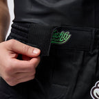 Close-up of a person wearing black martial arts pants with an elastic belt and a hook-and-loop closure. A hand is pulling the tab to demonstrate the closure.