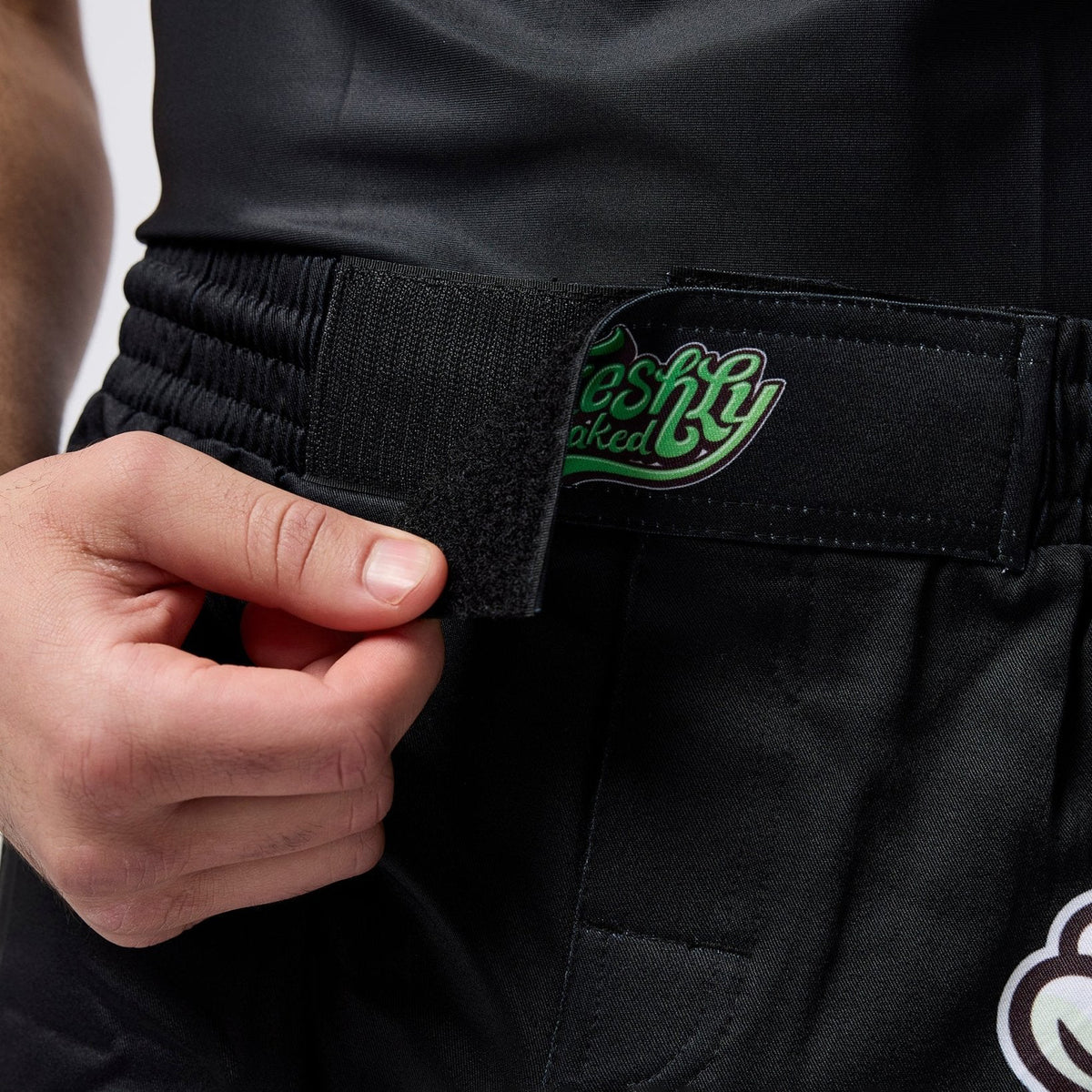 Close-up of a person wearing black martial arts pants with an elastic belt and a hook-and-loop closure. A hand is pulling the tab to demonstrate the closure.