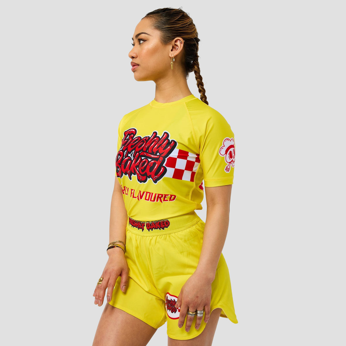 A woman modeling a bright yellow two-piece athletic set consisting of a short-sleeve t-shirt and matching shorts, featuring red and black graphic text on the front and a red and white checkered pattern on the sleeve.